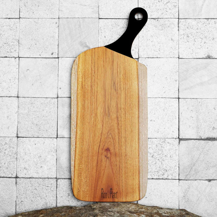 Teak cutting board with black handle displayed vertically on a tiled surface