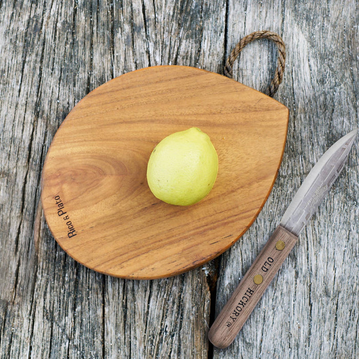 Rico & Plato limone teak cutting board serving platter with a rope handle and a fruit sitting on top of the platter next to a knife displayed on a wooden surface