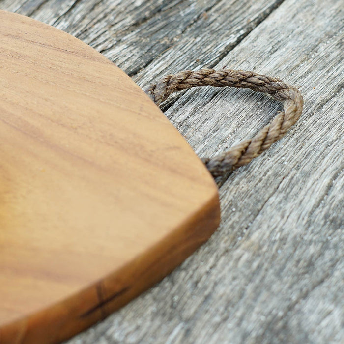 Close up of an irregularly shaped teak cutting board with a natural rope loop on one end, placed on a wooden surface.