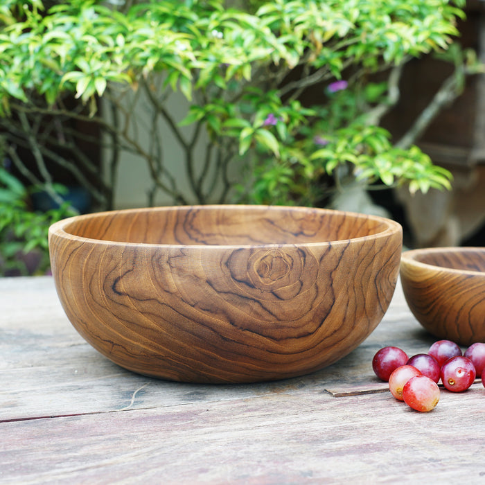 Large teak bowl next to a smaller teak bowl and grapes placed on a wooden surface with a nature background