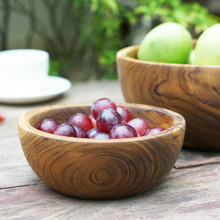 Small teak bowl filled with grapes and large teak bowl filled with apples both displayed outside on a wooden surface