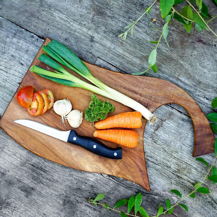 Top view of teak cutting board with curved handle with green onions, tomatoes, garlic, carrots, herbs, and knife displayed on a wooden surface
