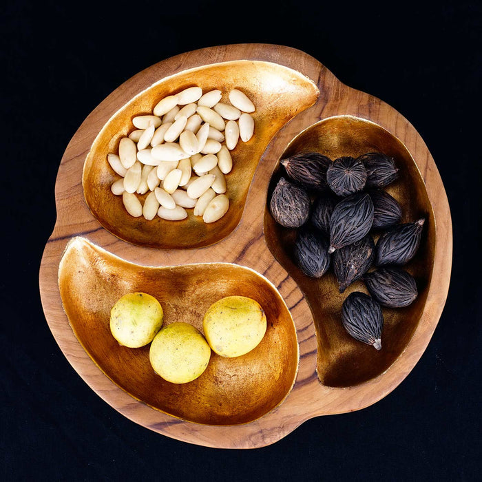 A teak wood serving bowl with a gold leaf interior, containing various food items like nuts and fruits, displayed on a dark background.