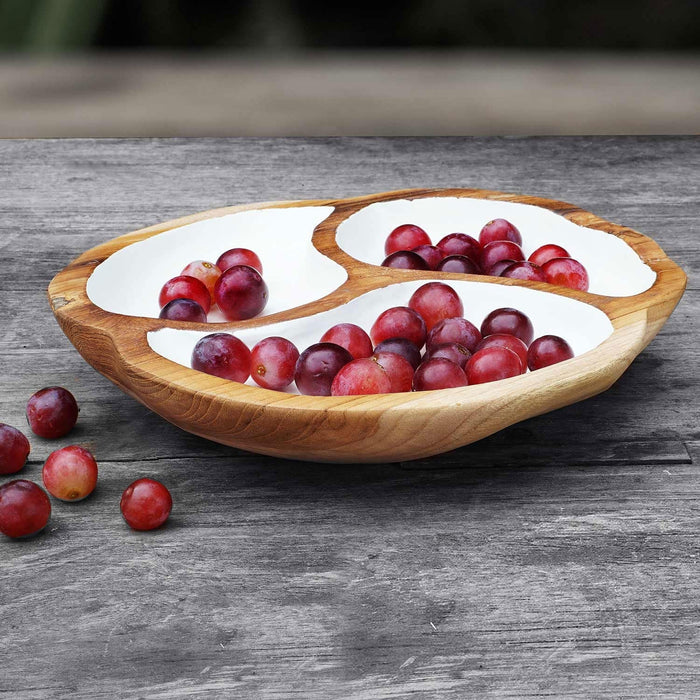 A hand-carved round Teak wood serving bowl with a white interior containing grapes, displayed on a wooden surface.