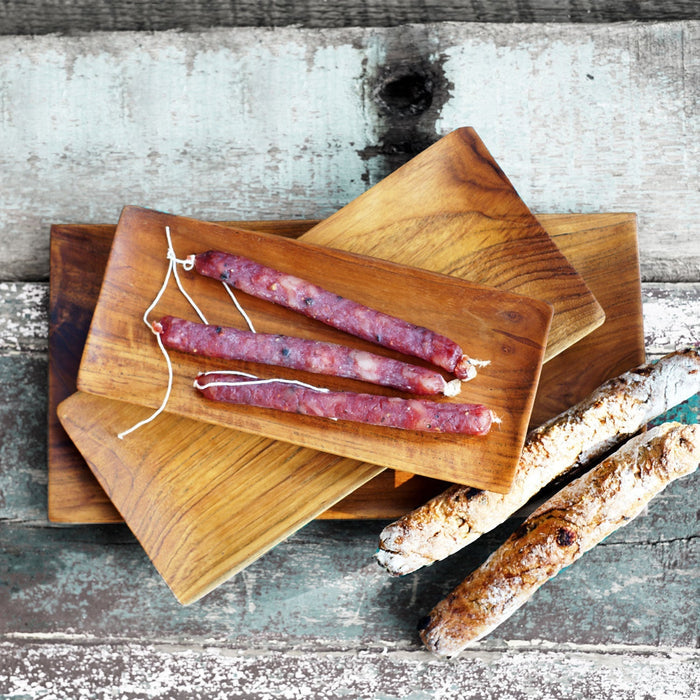 Top view of set of 3 Rico & Plato margot teak serving tray set with 3 sausage links on top with 2 breads on the side displayed on a whitewashed wooden surface