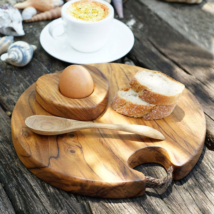 Teak cutting board with rope handle with egg, bread, wooden spoon, and cup of coffee displayed on a wooden surface