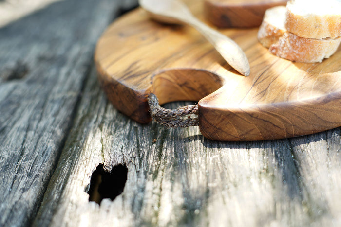 Close up of rope loop of teak cutting board with bread and spoon displayed on a wooden surface