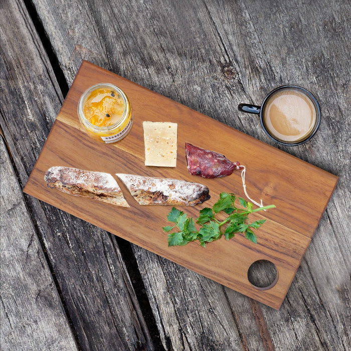 Top view of rectangle teak cutting board/serving platter with jam, cheese, bread, and sausage next to a cup of coffee displayed on a wooden surface