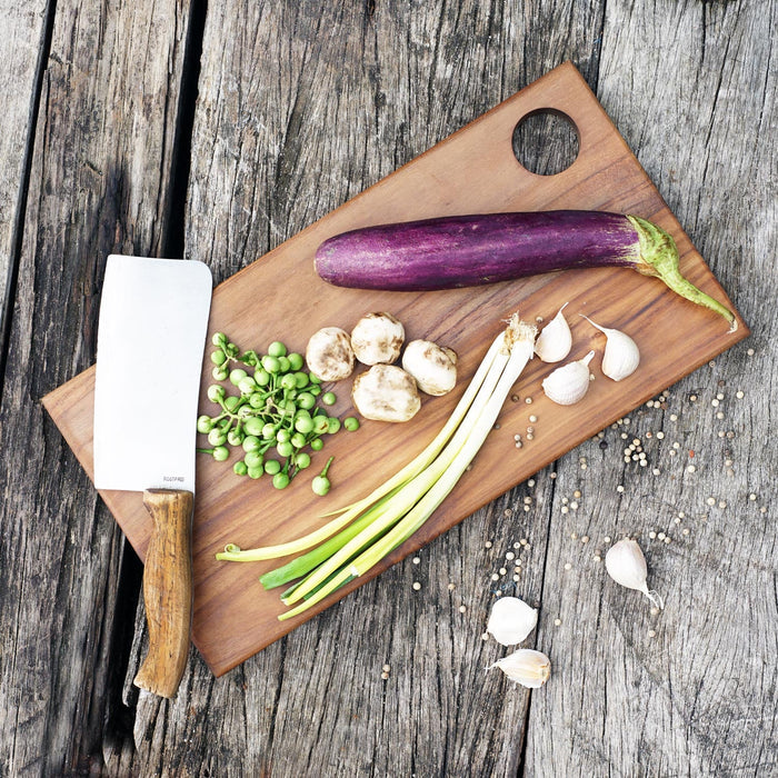 Top view of rectangle shaped teak cutting board/serving platter with green onions, mushrooms, eggplant, and garlic next to a knife displayed on a wooden surface