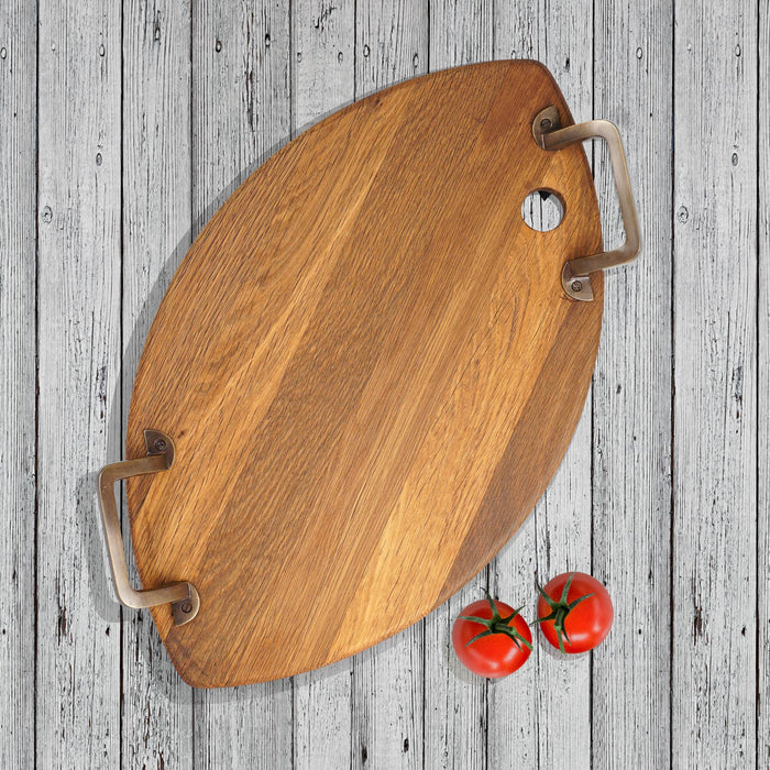Top view of oval shaped oak serving platter with metal handles next to tomatoes displayed on a white, wooden surface