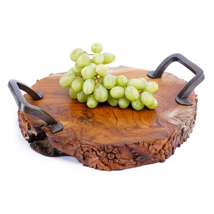 Teak serving platter with metal handles with decorations carved on the side of the serving platter with grapes on top displayed on a white background