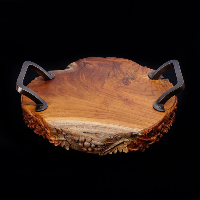 Teak serving platter with metal handles with decorations carved on the side of the serving platter displayed on a black background