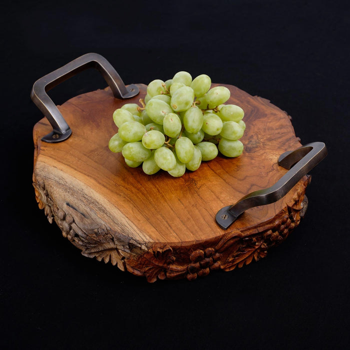 Teak serving platter with metal handles with decorations carved on the side of the serving platter with grapes on top displayed on a black background