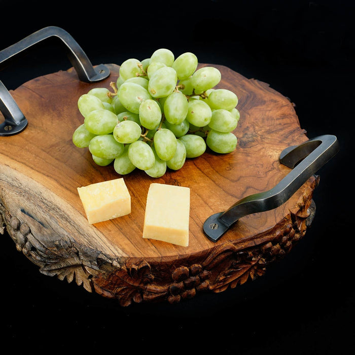 Teak serving platter with metal handles with decorations carved on the side of the serving platter with grapes and cheese on top displayed on a black background