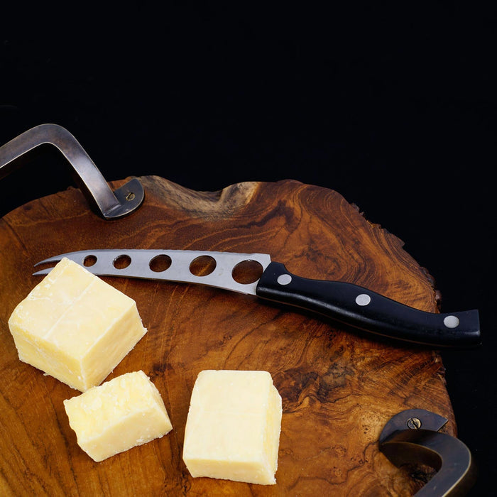 Close up of carved edge of teak serving platter with metal handles with cheese blocks and cheese knife displayed on a black background