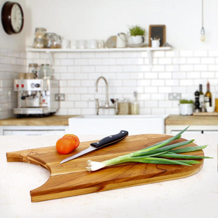 Teak cutting board with a plate cut out with a knife and some green herbs on it, displayed on a kitchen countertop.