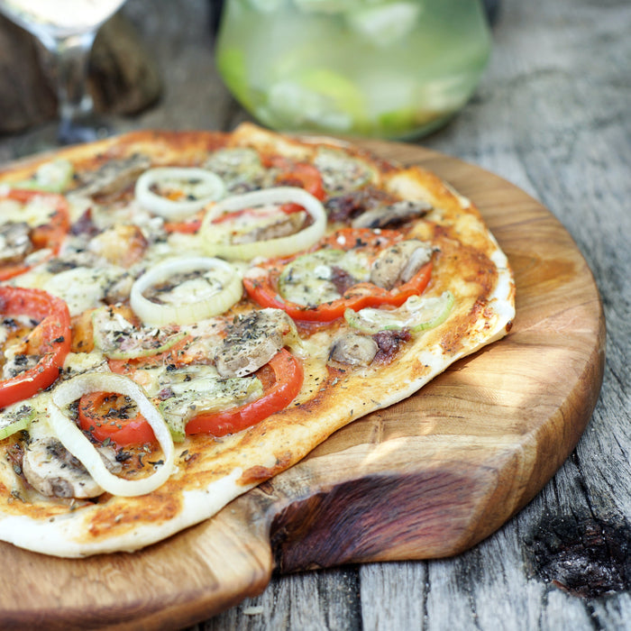 A teak wood pizza board with a pizza topped with tomato, onion, and mushroom slices, placed on a wooden surface.