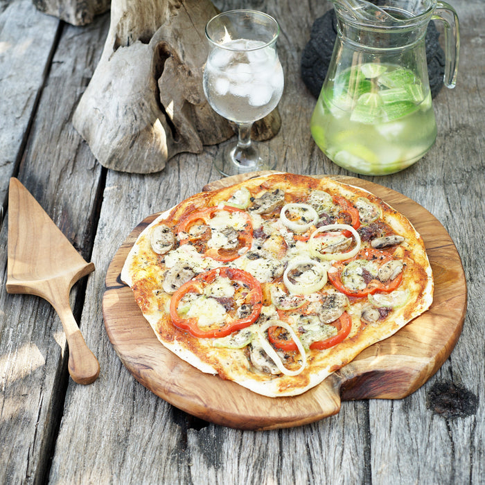 Teak pizza board with pizza placed on top next to a teak pizza serving spatula with water containers in the background displayed on a wooden surface