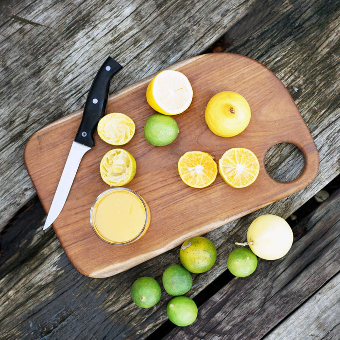 Top view of Rico & Plato olive teak cutting board serving platter with sliced lemons and limes next to a knife displayed on a wooden surface