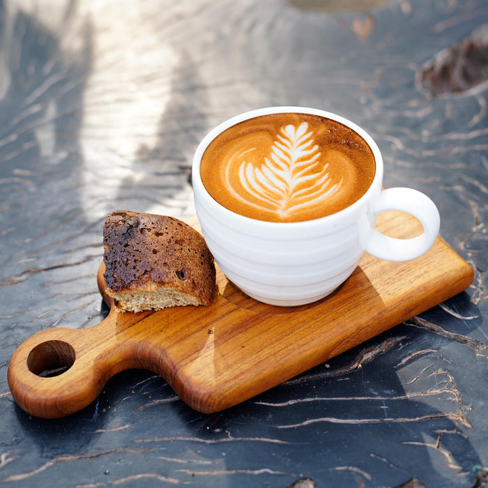 Teak coffee cup server with a mug of creamy coffee and slice of bread displayed on a black wooden countertop