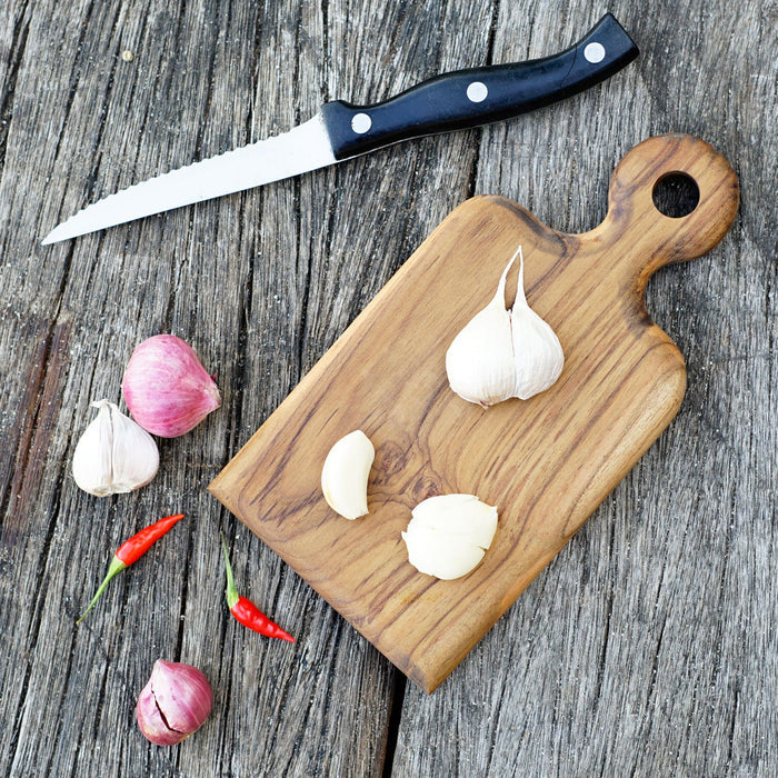 A teak cutting board with a handle, displaying garlic cloves, a knife, and red chili peppers on a wooden surface.