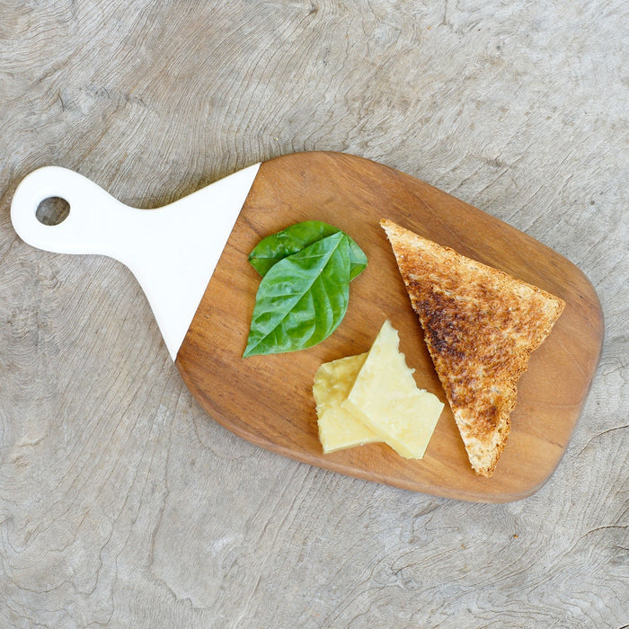 Top view of teak cutting board with white handle with toast, cheese, and herbs displayed on a wooden surface