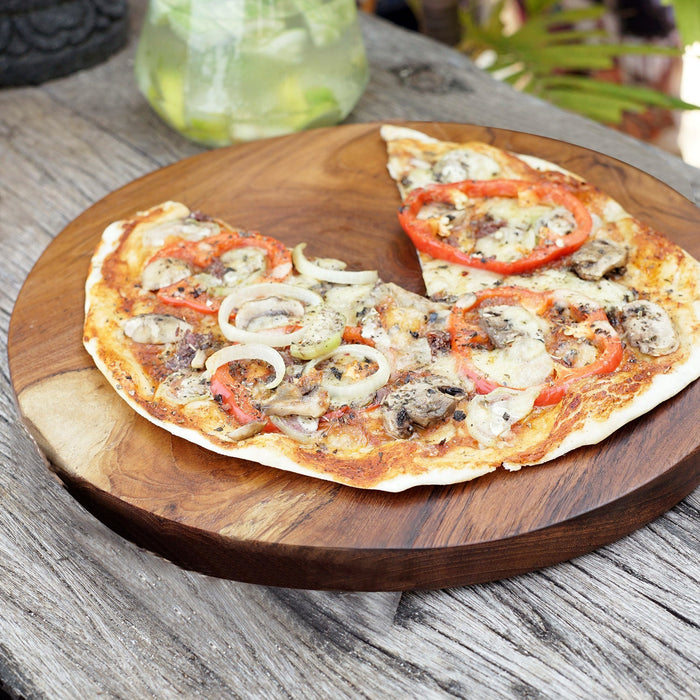 A teak wood pizza serving board with a pizza on top, placed on a wooden surface.