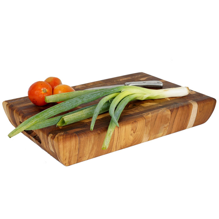 Rectangular end grain teak cutting board with knife, tomatoes, and green onions on top displayed on a white background