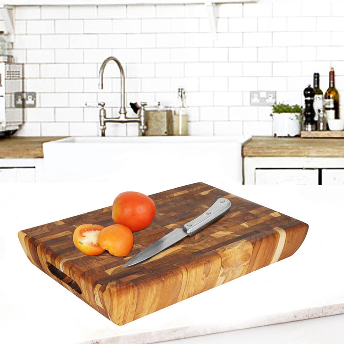 Rectangular end grain teak cutting board with carved out handle with a knife and tomatoes placed on top displayed in a kitchen setting