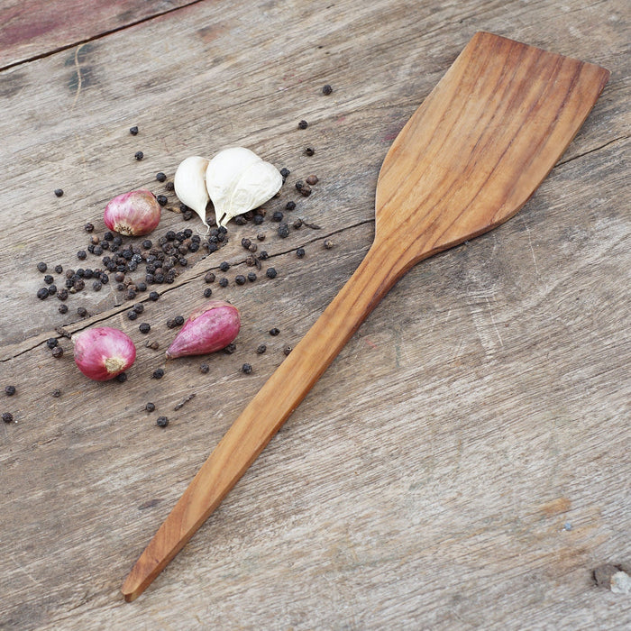 Extra large teak spatula next to shallot, garlic, and peppercorn displayed on a wooden surface