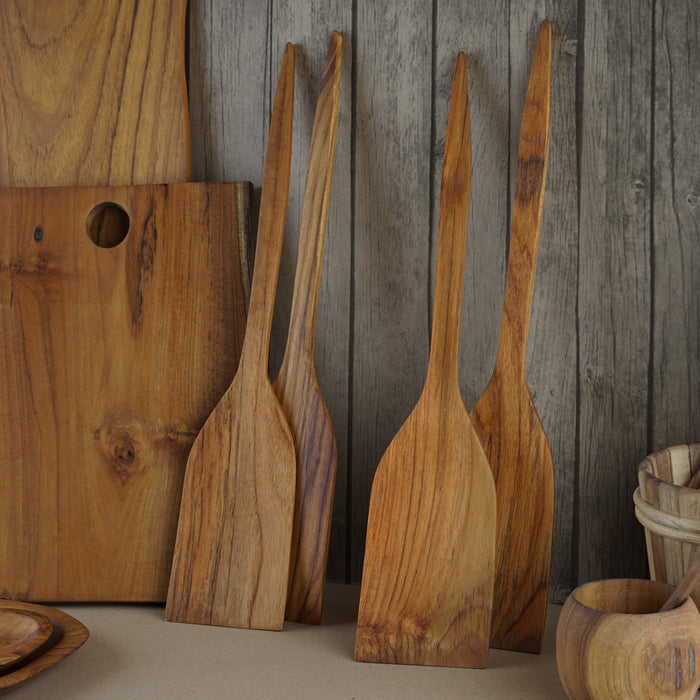 4 teak extra large teak spatula displayed vertically next to wooden cooking materials displayed on a wooden background