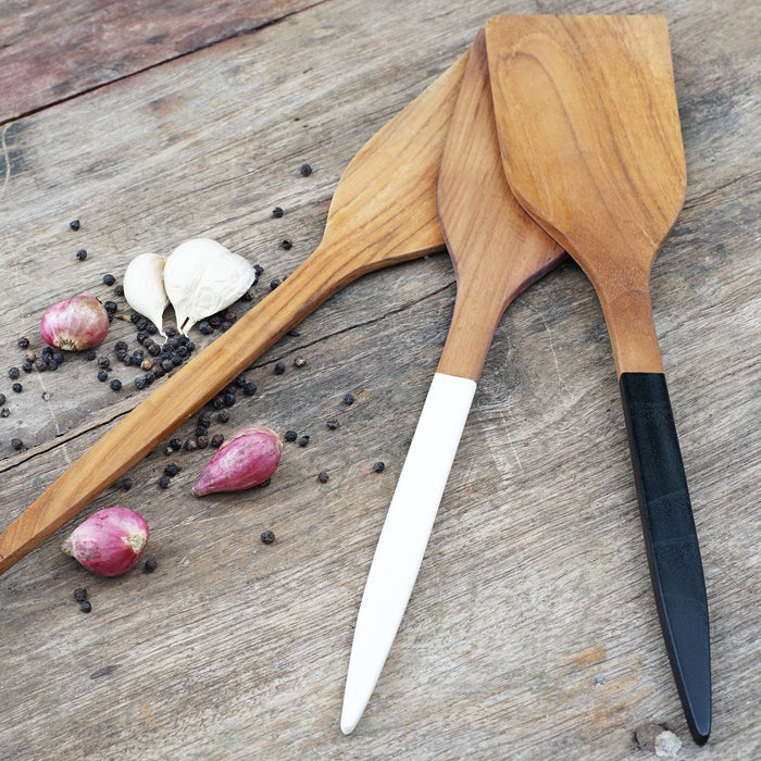 3 extra large teak spatula with natural handle, white handle, and black handle next to shallot, garlic, and peppercorn displayed on a wooden surface
