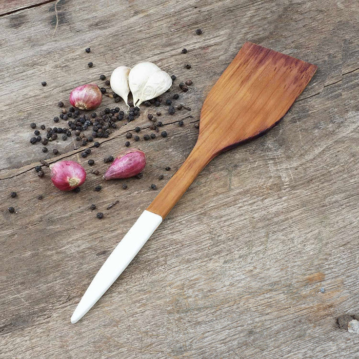 Rico & Plato resto teak spatula with white handle next to garlic, shallots, and peppercorns on a wooden surface
