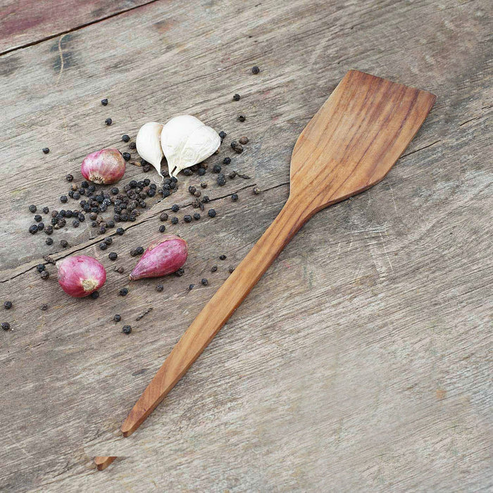 Teak spatula placed next to peppercorns, shallots, and garlic displayed on a wooden surface