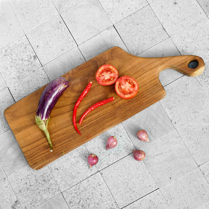 Top view of teak cutting board with natural teak handle with eggplant, chili, tomatoes, and shallots displayed on a tiled surface