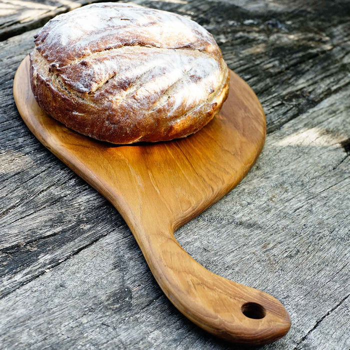 A paddle-shaped, teak wood cutting board with a smooth handle, displayed on a rustic wooden surface with a loaf of bread on top.
