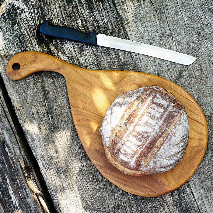 Rico & Plato savory teak cutting board serving platter with a circular bread on top of platter next to a bread knife displayed on a wooden surface
