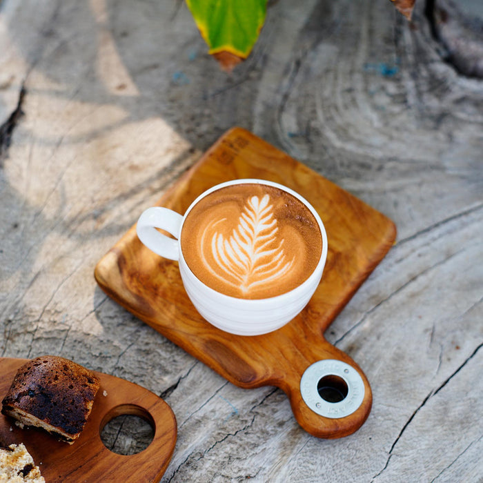 A rectangular teak wood coffee cup server with a white coffee cup placed on top, featuring a latte art design, served on a wooden surface.