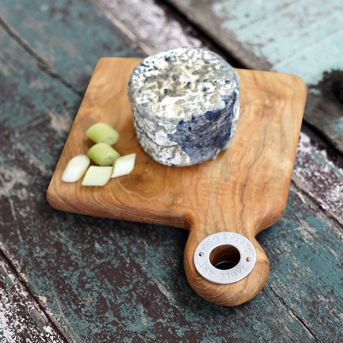 Squared shaped teak cutting board with a handle and cheese on top displayed on a wooden surface