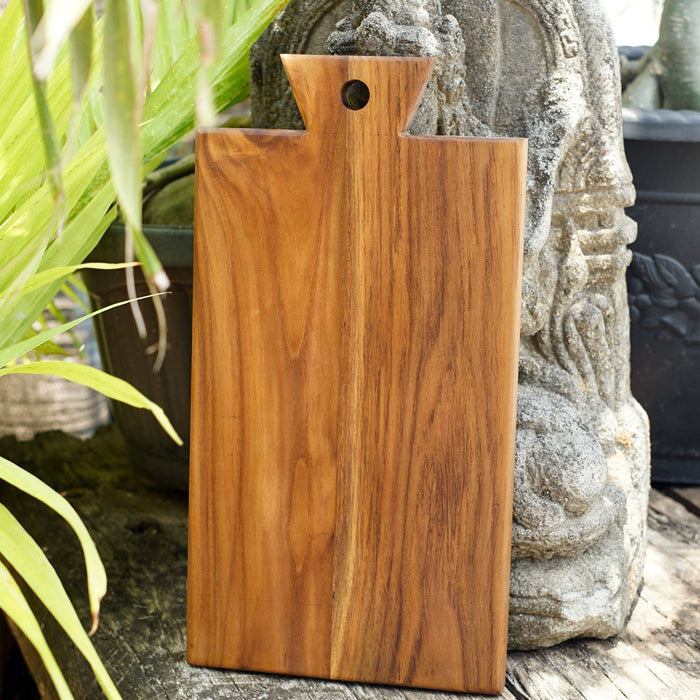 Front view of teak cutting board displayed vertically against a stone outside next to plants
