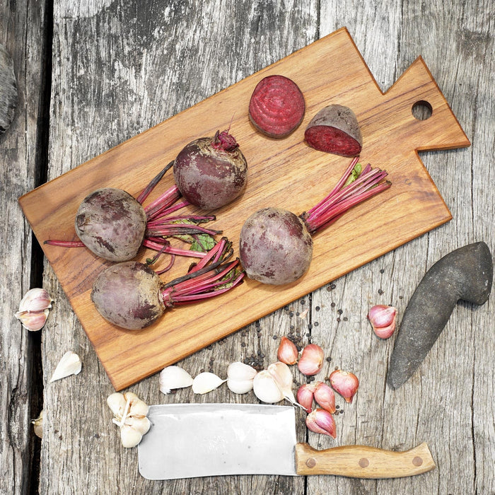 Top view of teak cutting board/serving platter with beets next to knife, garlic, and shallots displayed on a wooden surface