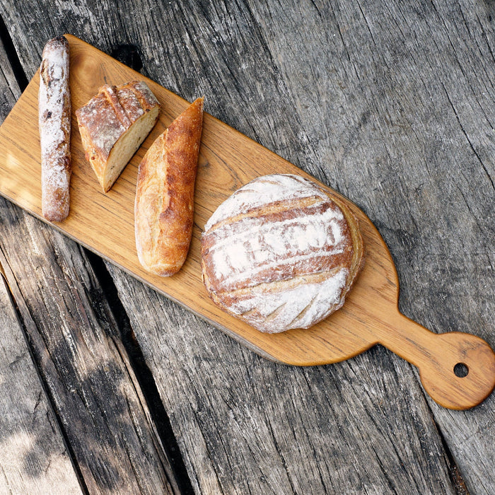 Rico & Plato sorrel teak cutting board serving platter with an assortment of bread on top displayed on a wooden surface