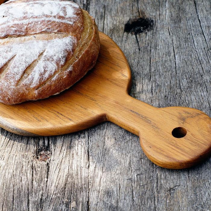 Close up of handle of Rico & Plato sorrel teak cutting board serving platter with bread sitting on platter displayed on a wooden surface