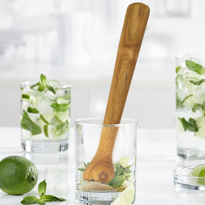 Teak cocktail tamper placed inside a clear shot glass with herbs and limes displayed on a white countertop with cocktails in the background