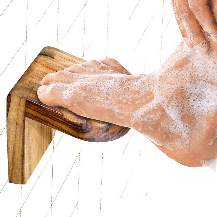 A person's foot with foam on it resting on a solid teak wooden shower footrest, placed on a white tiled wall.