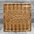 A square, brown teak wood cutting board with juice grooves and handles, crafted from durable end grain construction on a wooden background