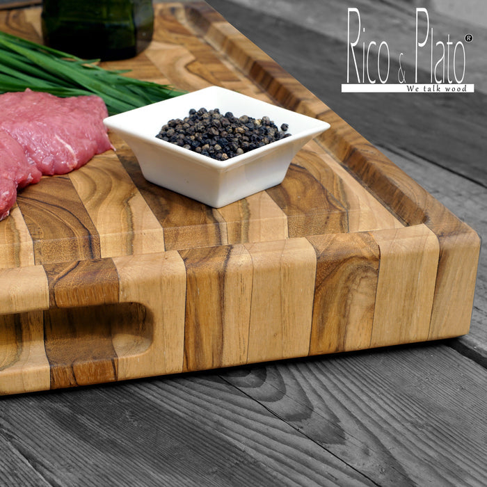 End grain cutting board with juice groove and handles with meat and chives on the board placed by a white dish with peppercorns on a wooden background