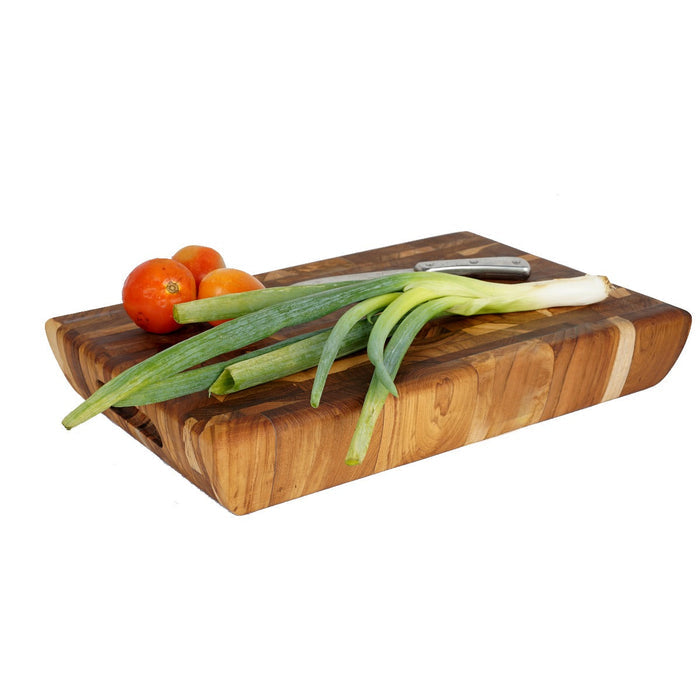 Rectangular end grain teak cutting board with knife, tomatoes, and green onions on top displayed on a white background