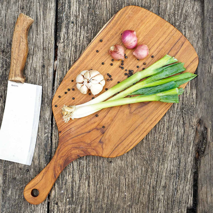 Top view of teak cutting board/serving platter shaped as a paddle with garlic, chives, shallots next to a knife displayed on a wooden surface