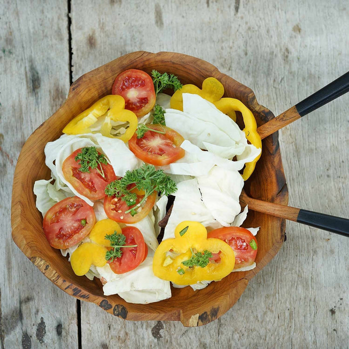 Teak salad fruit bowl with fruit and veggies inside with two serving utensils on a wooden background